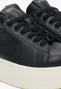 Zwarte leren sneakers hebben een ronde neus, een dikke witte tussenzool en platte zwarte veters door ronde oogjes. Smooth textuur met perforaties.