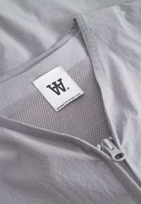 Lichtgrijs kledingstuk met een rits aan de voorkant, textuurstof en mesh-detail. Het label toont het merk “DOUBLE BY WOOD WOOD”.