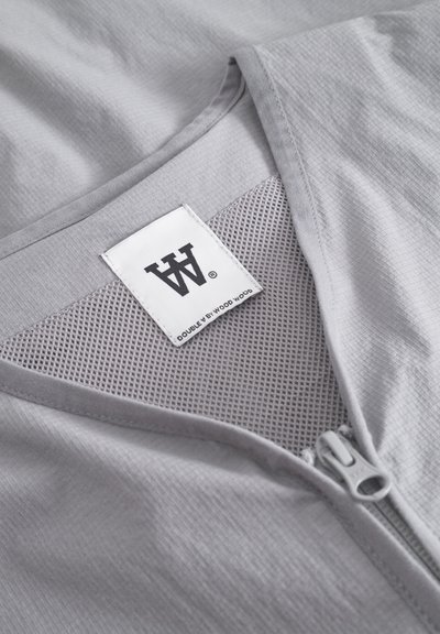 Vêtement gris clair avec une fermeture éclair à l'avant, un tissu texturé et des détails en maille. L'étiquette affiche le logo "DOUBLE BY WOOD WOOD".