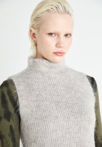 Maglioncino a collo alto grigio chiaro in maglia, con una consistenza morbida, caratterizzato da un design aderente e aperture delle maniche ampie, indossato sopra a una maglietta verde oliva.