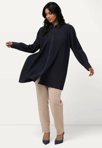Camicia a maniche lunghe blu navy con pieghe e colletto, abbinata a pantaloni beige chiaro e tacchi blu. Tessuto morbido, vestibilità rilassata.