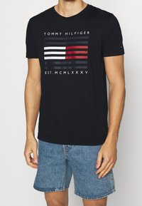 Camiseta de algodón negra con rayas horizontales en rojo, blanco y azul marino, con el texto "TOMMY HILFIGER EST. MCMLXXXV" debajo del diseño.
