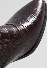 Närbild av en mörkbrun, glansig krokodilsmönstrad läderstövel med spetsig tå och detaljerad sömnad längs sömmen.