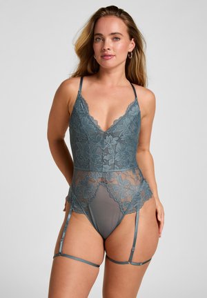 Frau trägt blau-grauen Spitzenbodysuit mit Blumenmuster und verstellbaren Strapshaltern vor neutralem Hintergrund.