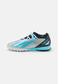 Zapato de fútbol con un empeine plateado, talón negro y acentos turquesas. Presenta una superficie texturizada, cordones y tacos de goma moldeada en la suela.