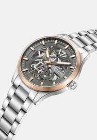 Zegarek skeleton z szarym cyferblatem, różowymi akcentami, stalową kopertą i bransoletą ze srebrnej stali nierdzewnej, prezentujący skomplikowane mechanizmy wewnętrzne.