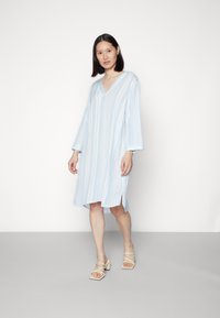 Vestido riscado em azul claro com decote em V, mangas longas e fendas laterais, feito de um tecido macio. Combinado com sandálias de salto bloco em bege.