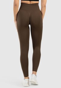Bruine high-waisted leggings van rekbaar materiaal, met een slim-fit ontwerp dat taper naar de enkels. Draagt met witte sneakers.