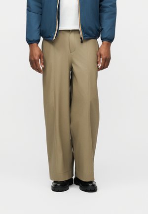 Moški, ki nosi bež široke hlače, črne robustne čevlje, belo srajco in modro prešito jakno z rokami sproščeno ob straneh.