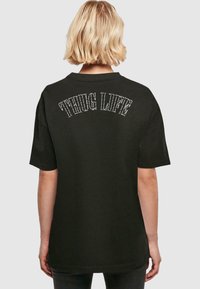 Juodas medvilninis marškinėlis su užpakalinėmis detalėmis, kuriose rausvai užrašyta "THUG LIFE". Trumpomis rankovėmis ir laisvu kirpimu.