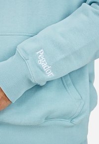 Felpa leggera azzurra con polsini e orlo a costine, con "Pegador" ricamato in bianco sulla manica sinistra. Materiale morbido con una texture liscia.