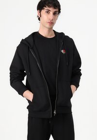 Sudadera negra con cremallera hecha de un tejido suave, con un bolsillo frontal y capucha con cordones. Presenta un diseño de rosa bordado en rojo y blanco en el pecho.