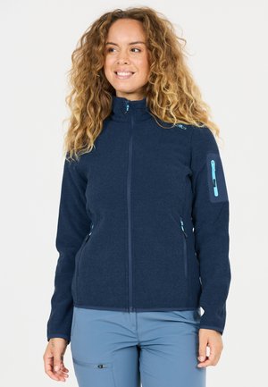 Kvinde med krøllet hår iført en marineblå jakke med lynlås og lyseblå bukser, smilende og vendt en smule mod venstre på en hvid baggrund.