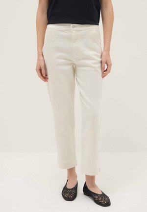 Mujer con pantalones rectos color blanco roto, top negro sin mangas y zapatos planos negros tejidos, de pie contra un fondo claro y liso.