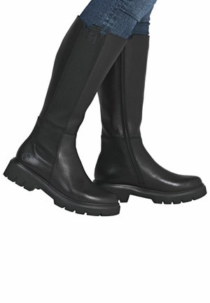 Bottes noires hautes en cuir lisse avec un panneau latéral élastique texturé. Comprend une semelle à crampons épaisse et une fermeture éclair sur le côté.
