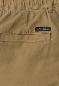 Rückseite der beigen Herrenhose mit elastischem Bund, einer Paspeltasche und einem schwarzen Etikett mit der Aufschrift "STREET ONE MEN Unmatched Comfort."