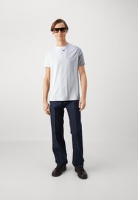 Camiseta blanca de manga corta con un pequeño logo, combinada con unos jeans de mezclilla oscuros con costuras azules y zapatos negros de cordones.