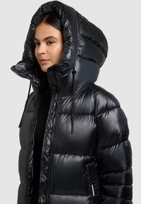 Chaqueta acolchada negra con cuello alto y capucha sobredimensionada. Fabricada con material brillante acolchado, con cordones ajustables y un bolsillo lateral.