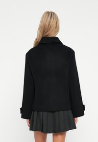 Plava žena u crnom vunenom sakou i sivoj plisiranoj suknji, stoji okrenuta leđima prema kameri na bijeloj pozadini.