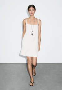 Weißes ärmelloses Kleid mit quadratischem Ausschnitt, strukturiertem Stoff, schlankem Design und lockerer Passform. Kombiniert mit dunkel gefärbten Flip-Flops und einer langen Halskette.