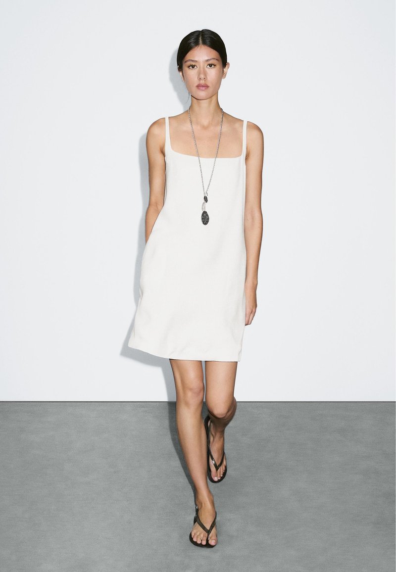 Weißes ärmelloses Kleid mit quadratischem Ausschnitt, strukturiertem Stoff, schlankem Design und lockerer Passform. Kombiniert mit dunkel gefärbten Flip-Flops und einer langen Halskette.