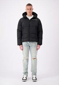 Zwarte pufferjas met capuchon, ritsluiting en twee voorzakken, gecombineerd met lichtblauwe distressed jeans en zwarte sneakers.