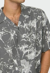 Kurzarmhemd aus dunkelgrauem Stoff mit großen weißen Blumenmustern, einem Eingeschnittenen Kragen und zwei Knöpfen vorne.