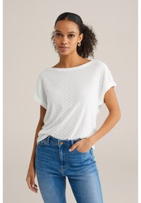 Balta trumpa rankovėmis palaidinė su tekstūruota žuvies kaulų struktūra, derinama su mėlynomis aukštomis džinsinėmis kelnėmis. Rankovės turi sukčių dizainą.