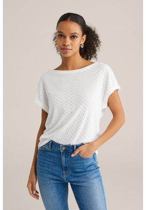 Fehér, rövid ujjú felső, amelynek textúrája halszálka mintázatú, kék, magas derekú farmernadrággal párosítva. Az ujjak felgöndörített kialakításúak.