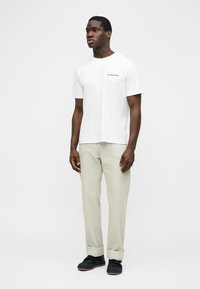 Bărbat stând, purtând un tricou alb cu mâneci scurte, pantaloni bej deschis cu tivuri și pantofi negri, pe un fundal simplu, deschis la culoare.