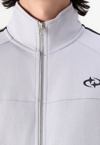 Lichtgrijze zip-up jas met een hoge kraag, voorzien van zwarte zijstrepen en een logo aan de linkerkant. Soepele stoftextuur.