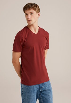 Tricou V cu guler de culoare bordeaux, realizat dintr-un material moale. Mâneci scurte, croială slim cu fante laterale. Purtați-l cu blugi de culoare albastru deschis, pe un fundal simplu.