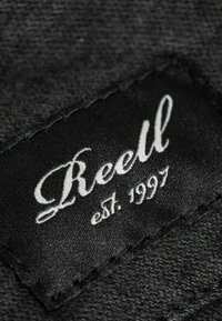 Black fabric label with white cursive text reading "Reell," established 1997, stitched at the edge. Textured fabric background.