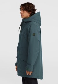 Tmavý teal parka kabát s kapucňou, s uzáverom na zips a gombíky, prednými vreckami a nastaviteľnou šnúrkou. Textúrovaná tkanina.