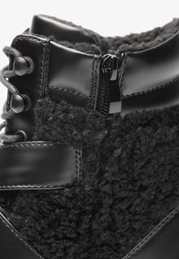 Fekete, magas szárú csizma, amely fényes fekete bőrből és texturált, bolyhos anyagból készült. Oldalsó cipzárral és kerek fekete fűzőkkel rendelkezik.