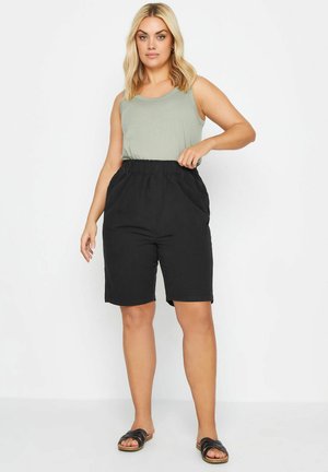 Kvinde iført grøn ærmeløs top, sorte knælange shorts med høj talje og sorte slid-sandaler, stående mod en hvid baggrund.