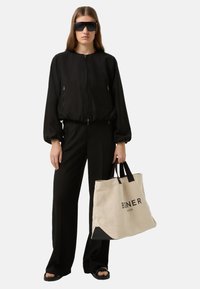 Czarna kurtka typu bomber z pełnym zamkiem, luźnym krojem, zestawiona z czarnymi spodniami i czarnymi sandałami. Torba na ramię z płótna z czarnymi akcentami i logo.
