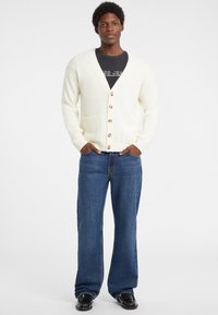 Crème gebreide cardigan met een V-hals, houten knopen en voorzakken, gecombineerd met blauwe denim jeans en zwarte schoenen.