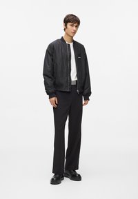 Jauns vīrietis stāv, tērpies melnā bomber jakā, baltā krekla, melnās biksēs, melnās kurpēs un pelēkā jostā pret vienkāršu baltu fona.