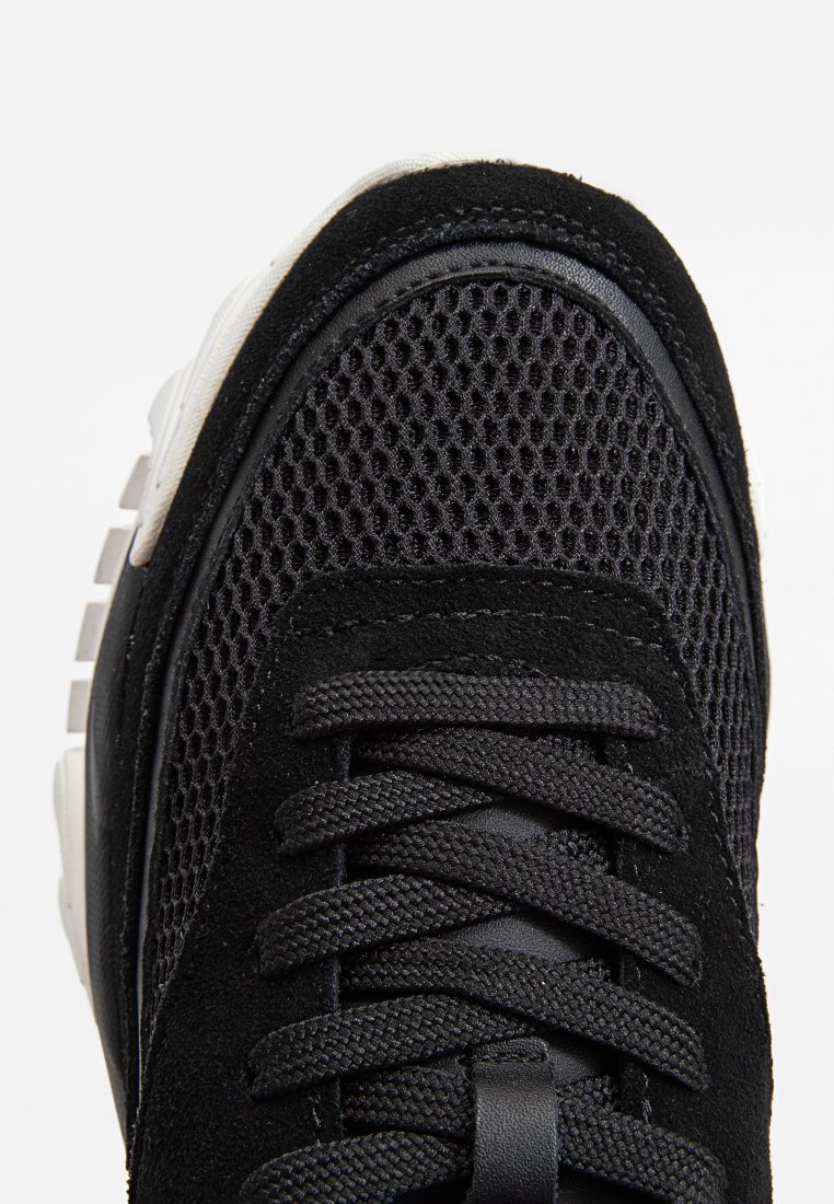Baskets noires avec une tige en mesh, des accents en daim, des lacets texturés noirs et une semelle blanche contrastante. Vue en gros plan du design.