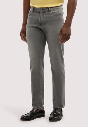 Uomo con jeans skinny grigi, mocassini in pelle nera e camicia gialla, in piedi contro uno sfondo chiaro e uniforme.