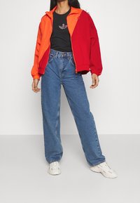 Person klädd i en röd och orange färgblockjacka, svart t-shirt med logga, höga lösa blå jeans och vita sneakers, stående mot en vit bakgrund.