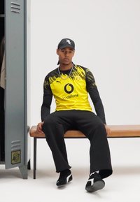 Geltonos spalvos „Puma“ sportinis marškinėlis su juodais akcentais, kuriame yra apskritas logotipas ir BVB ženklelis. Juodos kelnės ir sportbačiai užbaigia aprangą.