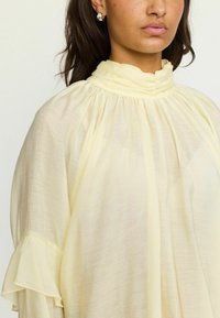 Blouse jaune clair avec un col haut, un tissu froncé et des manches fluides avec des volants. Fabriquée en matériau doux et transparent.