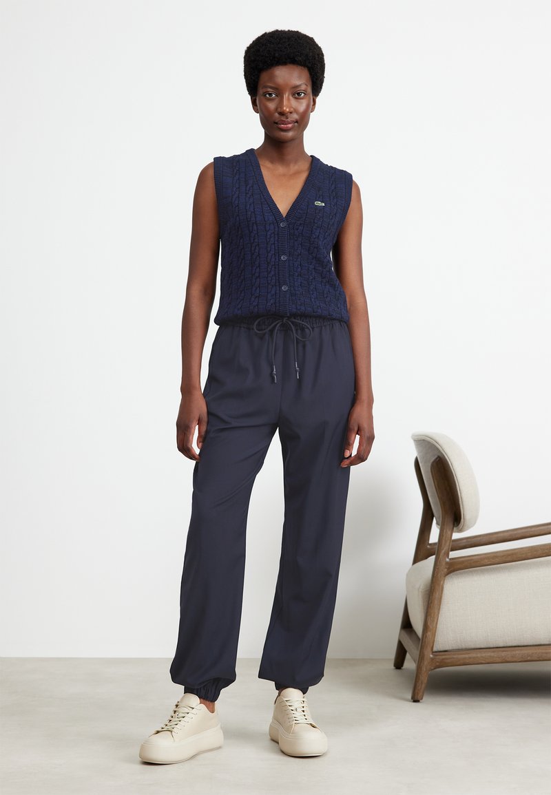 Granatowy dzianinowy kamizelka z wzorem warkocza i guzikami, w połączeniu z ciemnoszarymi spodniami elastycznymi w talii i beżowymi trampkami. Prosty design, swobodny krój.