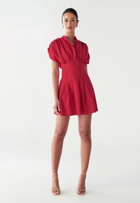Rotes kurzes Kleid mit tailliertem Oberteil, gepufften Ärmeln und plissiertem Rock. Aus glattem Stoff gefertigt. Kombiniert mit nude-farbenen Riemchen-Heels.