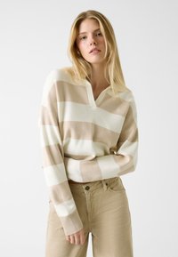 Pull en maille rayé beige et crème avec col en V et poignets côtelés, porté avec un pantalon beige clair, orné d'un logo brodé.