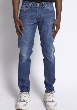 Man draagt strakke blauwe jeans en witte sneakers, staat met ontspannen handen langs zijn zij tegen een effen achtergrond.