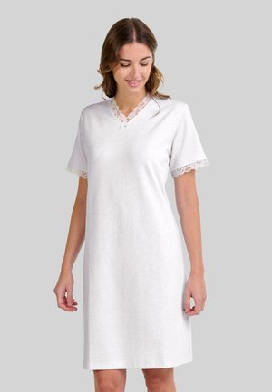 Mujer vestida con un camisón blanco de manga corta con encaje en el escote y las mangas, de pie frente a un fondo gris liso.