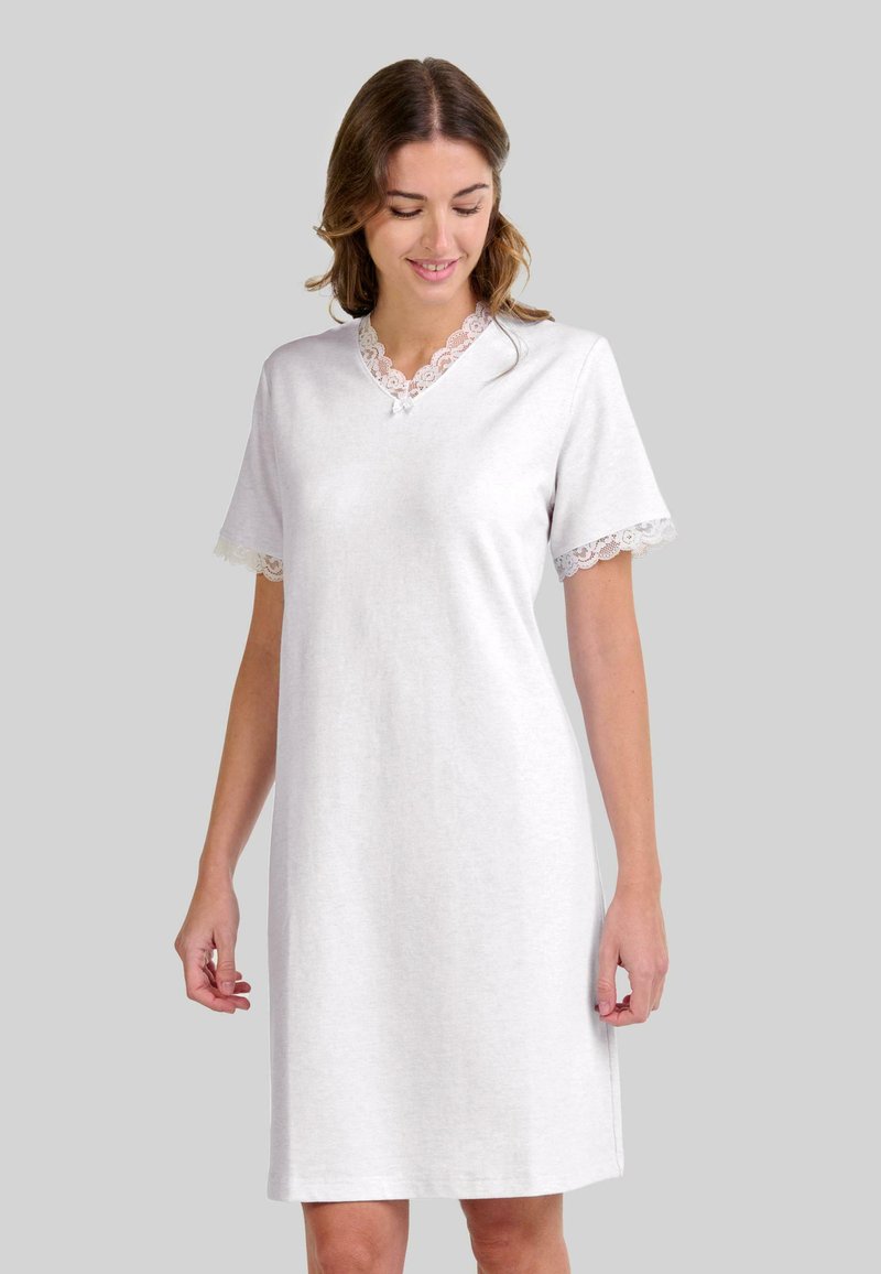 Mujer vestida con un camisón blanco de manga corta con encaje en el escote y las mangas, de pie frente a un fondo gris liso.
